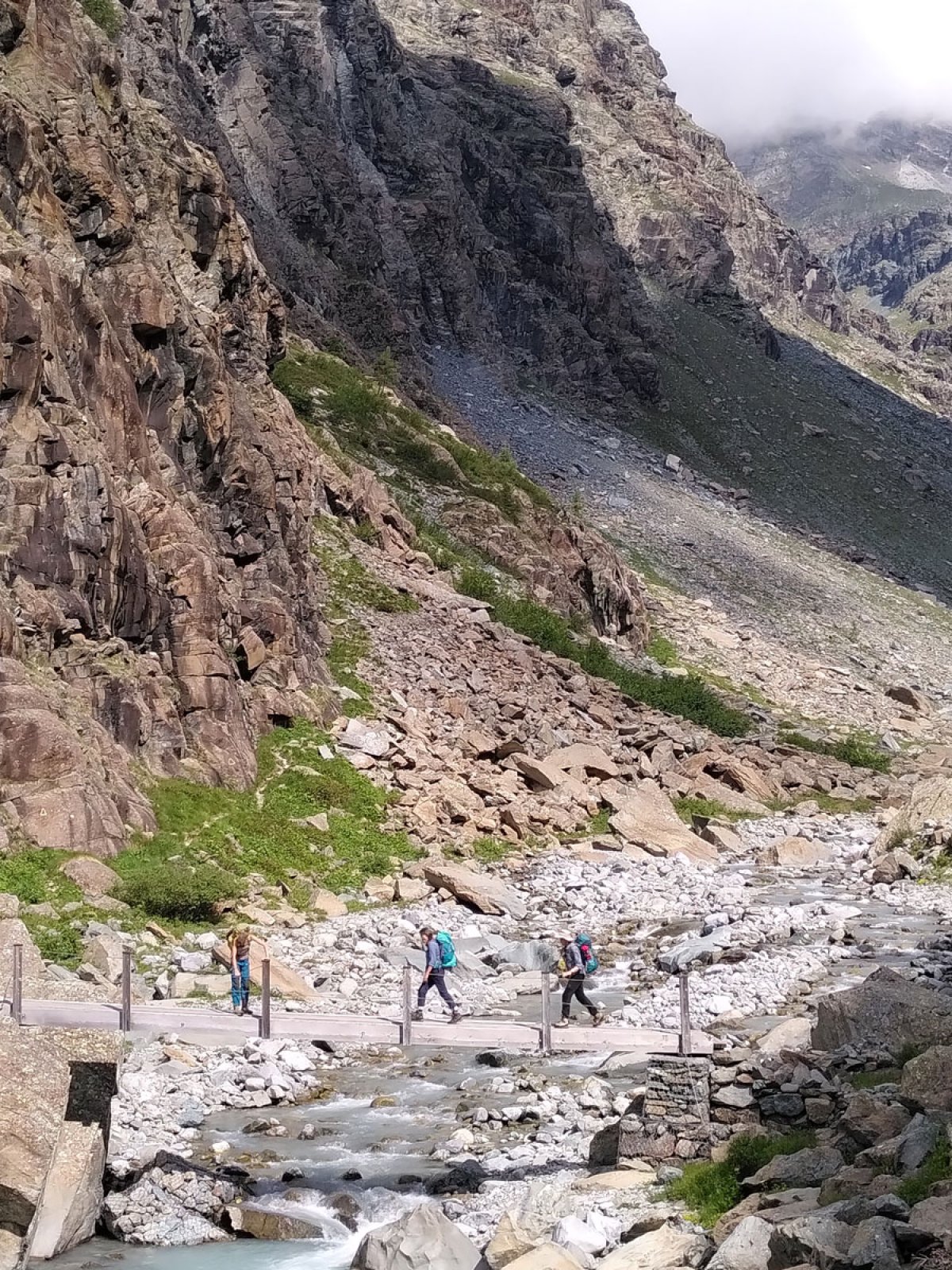 Drei Wanderinnen laufen über eine Brücke, hinter ihnen eine steile, bräunliche Felswand