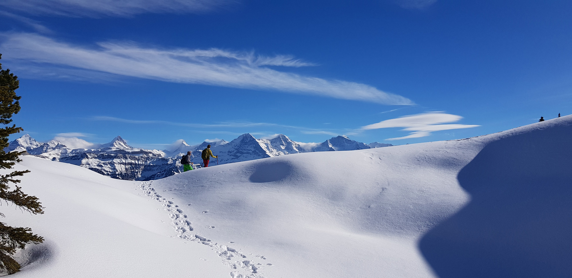 Zwei Personen wandern mit Schneeschuhen - im Hintergrund das Panorama der schneebedeckten Berner Alpen. 